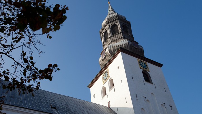 En høj kirkespire med et ur og en spir står mod en klar blå himmel.
