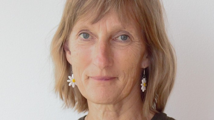 En kvinde med kort blondt hår iført en elegant brun top og fine øreringe poserer mod en ensfarvet baggrund.