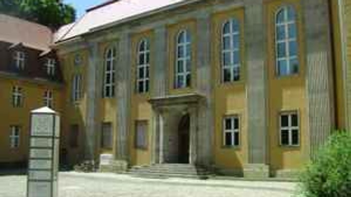 Historisches gelbes Gebäude mit Bogenfenstern und einem zentralen Eingang, umgeben von einem Kopfsteinpflasterhof.