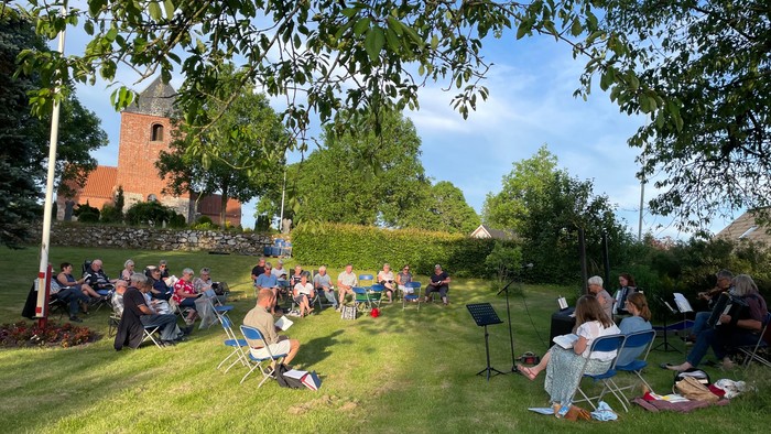En gruppe af mennesker sidder på græs og lytter til en koncert under træer ved en kirke.