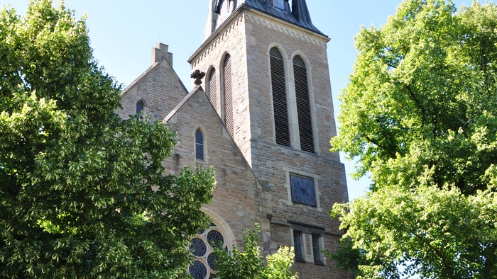 Historische Steinkirche mit hohem Turm und Bogenfenstern, umgeben von üppigen grünen Bäumen.