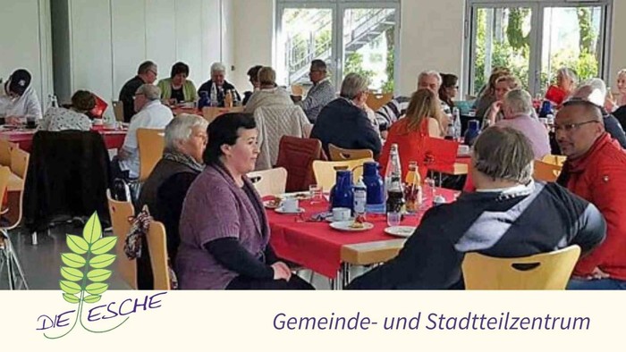 Gruppe von Menschen, die in einem Gemeinschaftszentrum bei einer Veranstaltung gemeinsam essen und sich unterhalten, mit roten Tischdecken und gelben Stühlen.