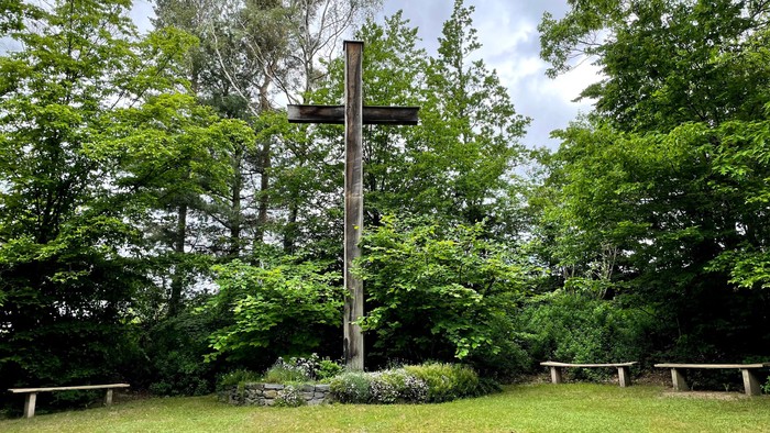 Ein hohes Holzkreuz steht in einem grünen Park mit Bänken.