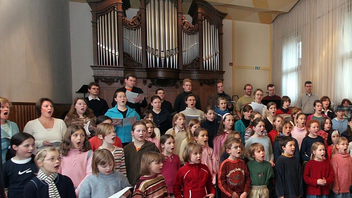 Eine vielfältige Gruppe von Kindern und Erwachsenen, die gemeinsam in einer Kirche singen, mit einer großen Orgel im Hintergrund.