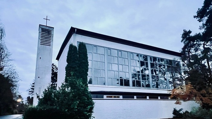 Moderne Kirchengebäude mit großen Glasfenstern und einem hohen Glockenturm neben üppigem Grün.