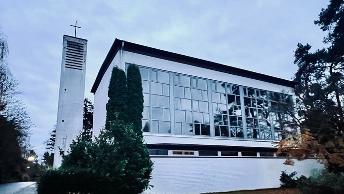 Moderne Kirchengebäude mit großen Glasfenstern und einem hohen Glockenturm neben üppigem Grün.