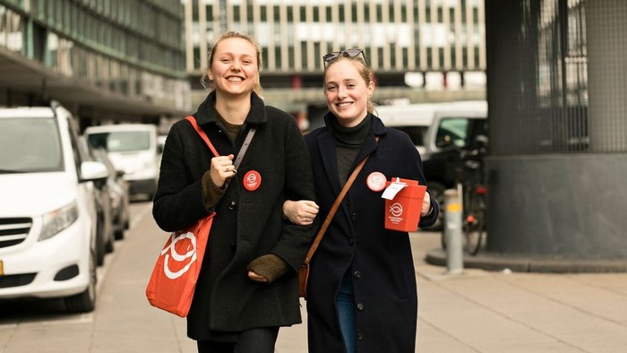 To glade personer går sammen og holder kaffekopper, mens de bærer tasker med røde logoer på en bygade.