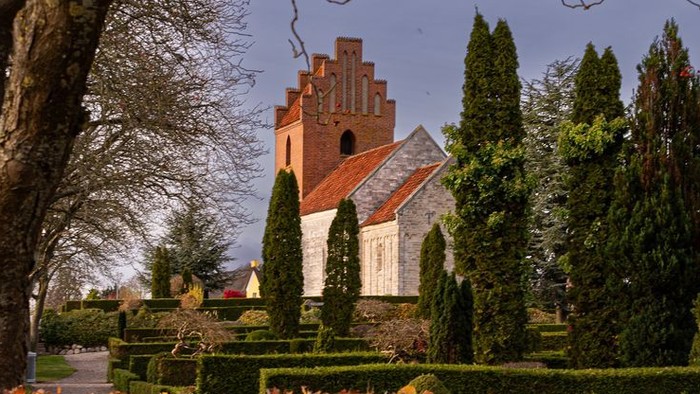 En gammel kirke med spiretårn omgivet af høje træer og efterårsblade.