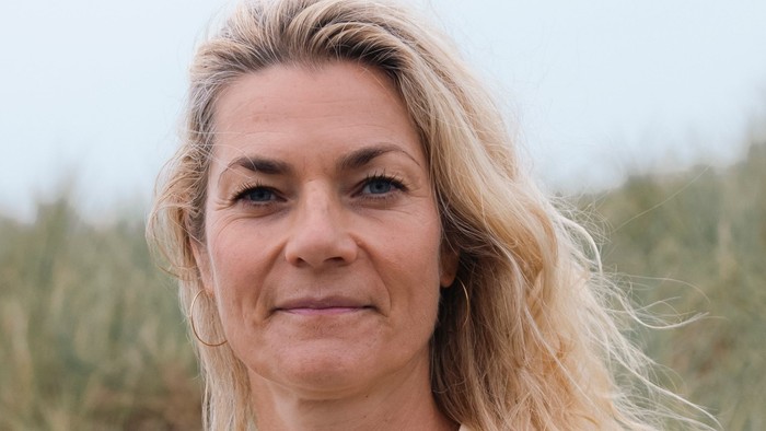 En kvinde med langt blondt hår i en lys top poser udendørs mod en naturlig baggrund.
