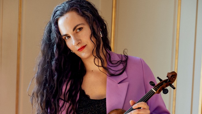 En kvinde i en lyserød jakke holder og poser med en violin foran en elegant baggrund.