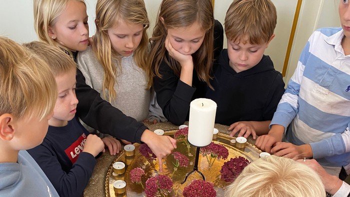 En gruppe børn samles omkring et bord oplyst af stearinlys med blomster, hvor de deltager i en ceremoni eller aktivitet.