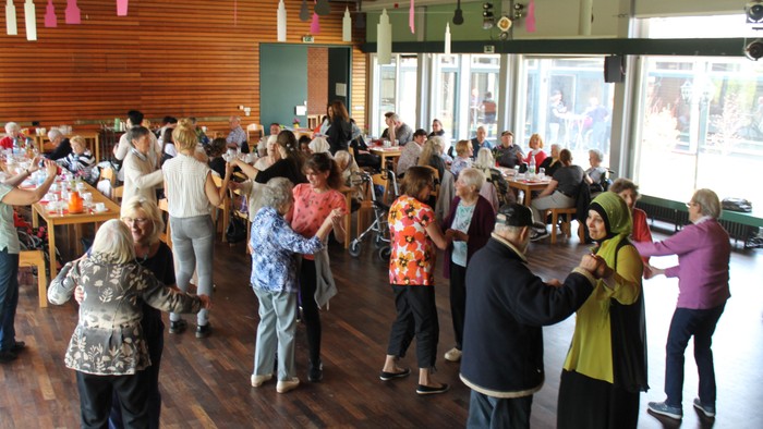 Eine Gruppe älterer Menschen tanzt in einem großen Raum mit Holzboden und großen Fenstern.