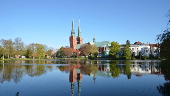 Kirche mit zwei Türmen, die sich im ruhigen Wasser spiegelt, umgeben von Gebäuden und Bäumen unter klarem blauem Himmel.