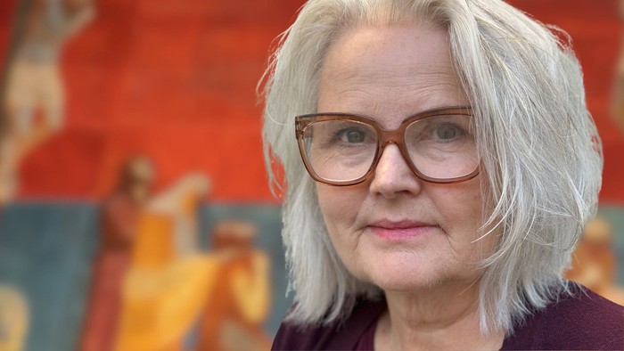 En kvinde med gråt hår og briller står foran en farverig, abstrakt muralbaggrund.