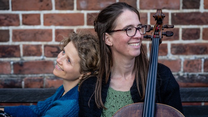To kvinder smiler, den ene holder en cello op ad en murstensvæg.
