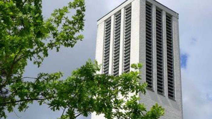 Moderner Kirchturm mit Kreuz, eingerahmt von üppigem grünem Laub unter einem bewölkten Himmel.