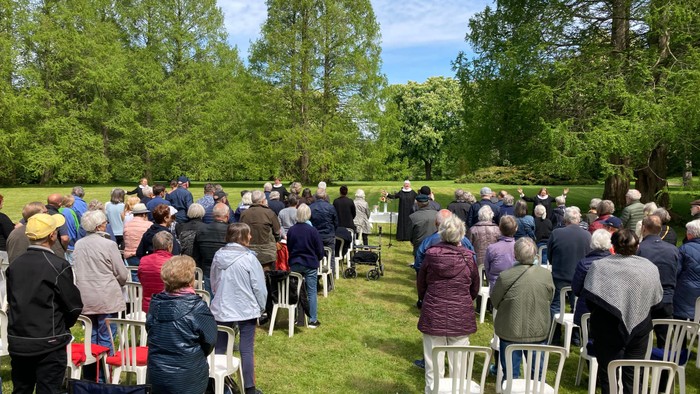 En stor gruppe af mennesker sidder på stoler i en park og ser mod en scene.