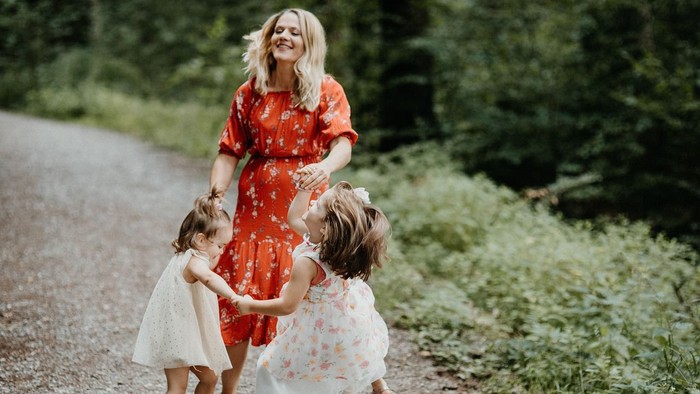 Eine Frau in einem roten Kleid geht Hand in Hand mit zwei jungen Mädchen in weißen Kleidern auf einem Feldweg.
