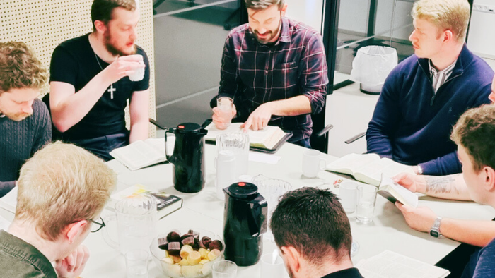 En gruppe mænd samlet omkring et bord, hvor de diskuterer med en åben Bibel og snacks til stede.