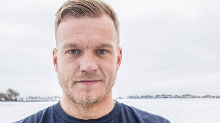 En mand med kort hår og skæg står udendørs ved et vand.