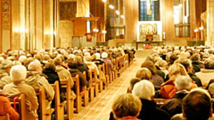 Eine große Gemeinde versammelt sich in einer prächtigen Kathedrale während eines Abendgottesdienstes, sitzend und aufmerksam.