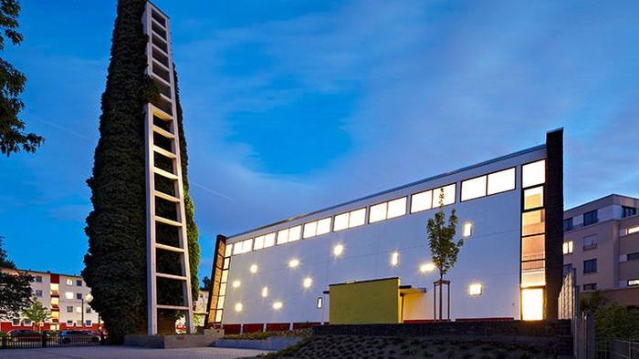 Moderner Gebäudekomplex, der bei Dämmerung beleuchtet ist, mit einer hohen vertikalen Struktur und angrenzender eleganter Architektur.