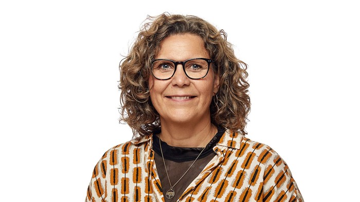 En glad person med krøllet hår, briller og en mønstret blazer, der smiler til kameraet.