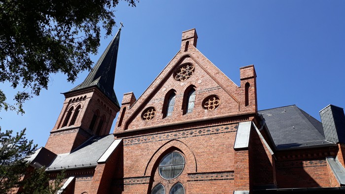 Das Bild zeigt eine rote Ziegelkirche mit spitzen Bögen und einem hohen Turm vor einem klaren blauen Himmel.