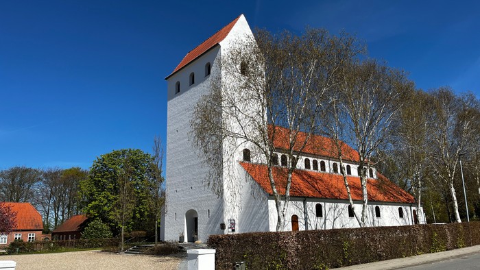 Historisk hvid kirke med rødt tegltag og et højt firkantet tårn omgivet af træer.