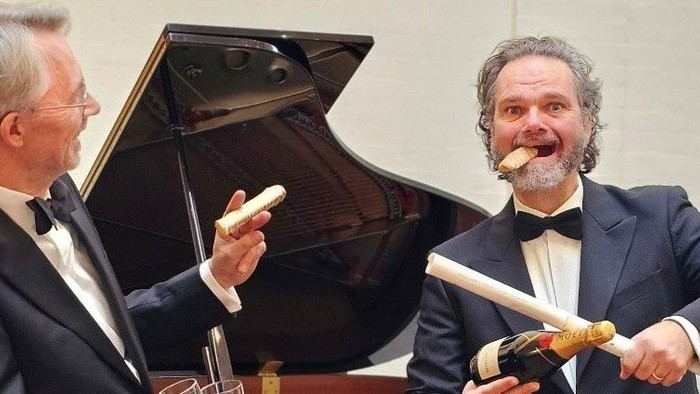 To mænd i jakkesæt spiller på instrumenter, den ene på et klaver.