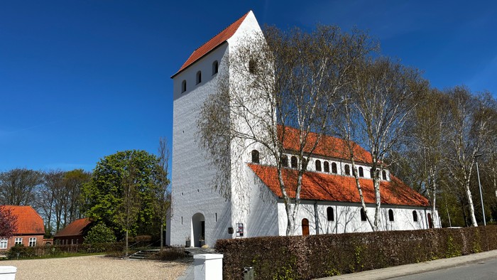 Historisk hvid kirke med rødt tegltag og et højt firkantet tårn omgivet af træer.