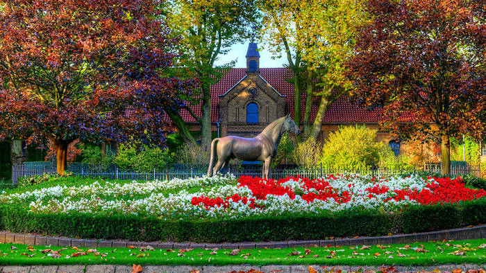 Ein Pferd steht in einem blühenden Garten vor einem Haus mit rotem Dach und umgeben von bunten Bäumen.