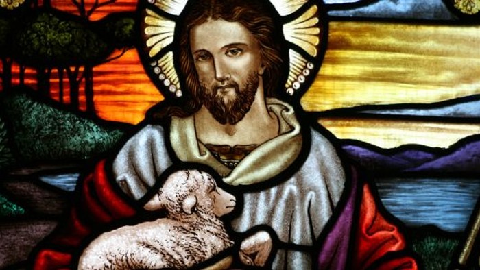 Glasmaleri af Jesus, der holder et lam, med en fredelig landskabsbaggrund.