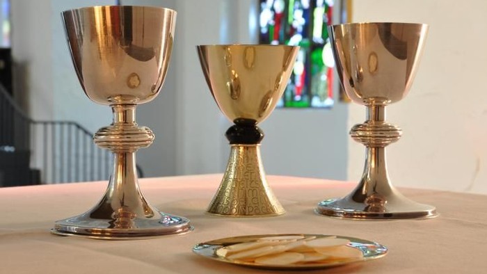 Drei verzierte Abendmahlskelche mit einer Hostie auf einer runden Platte, vor einem Hintergrund aus Buntglasfenstern.