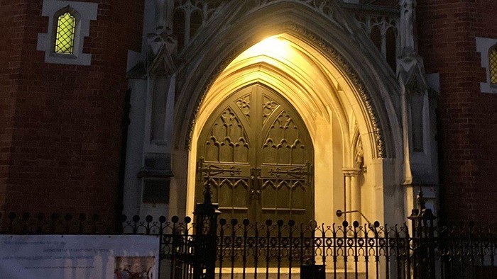 Gotischer Kirchenportal-Eingang, nachts beleuchtet, mit verzierten schmiedeeisernen Toren und Backsteinfassade.