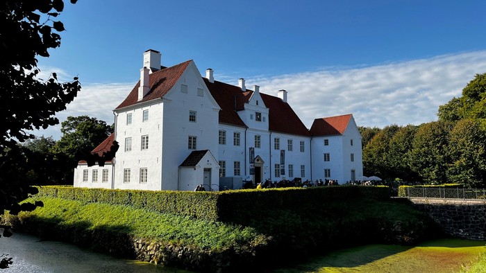 Et malerisk hvidt gods med røde tage ligger ved siden af en rolig, grøn skrænt og flydende vand.