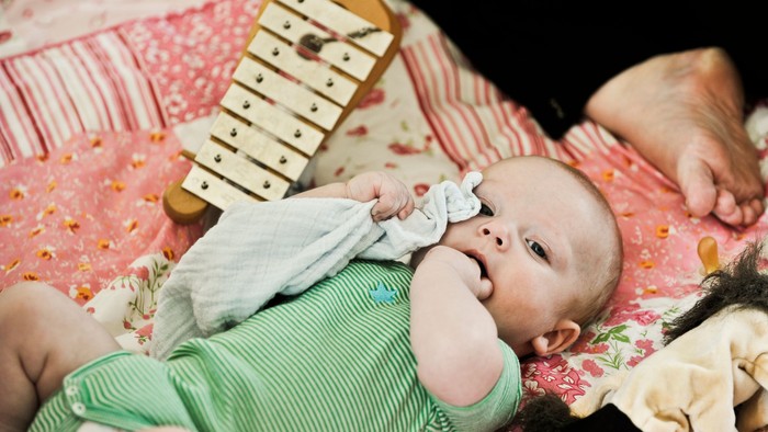 Et spæd barn ligger på en rosafarget dekke og ser opad, mens en voksende spiller på et xylofon.