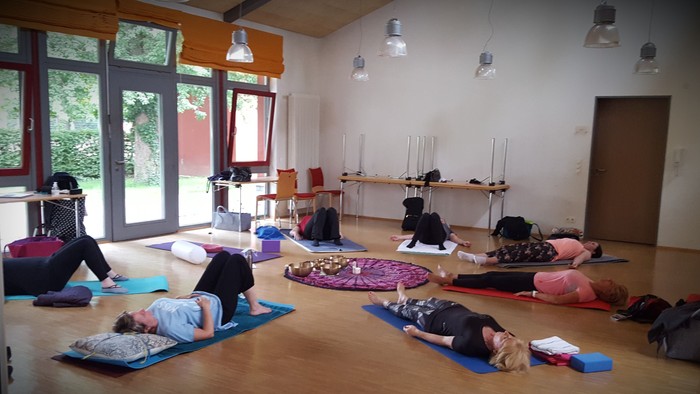 Yogaklasse in einem hellen Raum mit großen Fenstern und Holzboden.