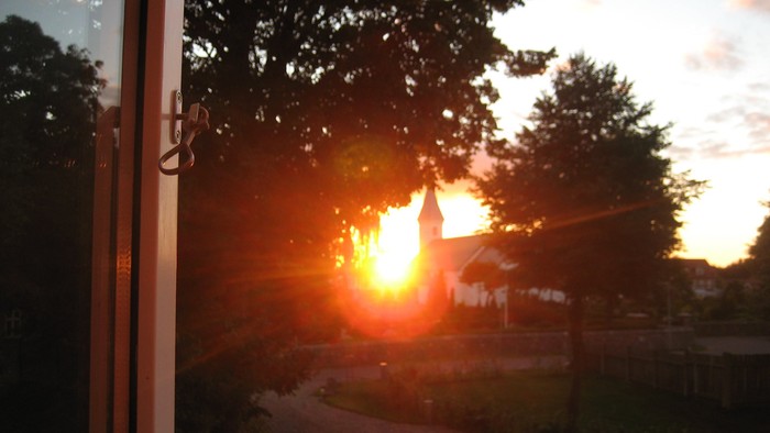 Solnedgang set over en vej med træer og en kirke i baggrunden