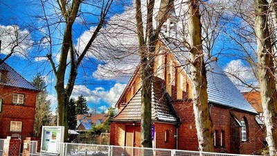 Ein rotes Backsteingebäude mit spitzem Dach und einem Kreuz darauf, umgeben von kahlen Bäumen und einem blauen Himmel.