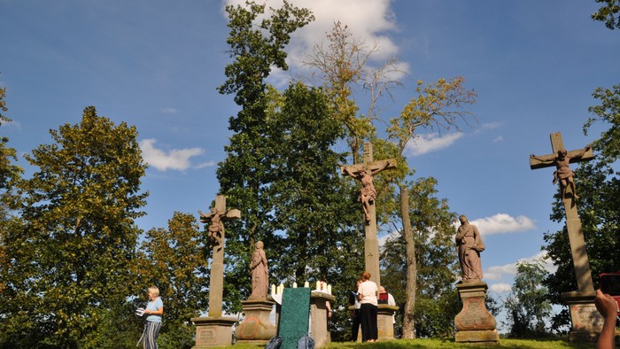 Einige Personen stehen auf einem grasbewachsenen Hügel mit mehreren Grabsteinen und Kreuzen umgeben von Bäumen unter einem klaren blauen Himmel.