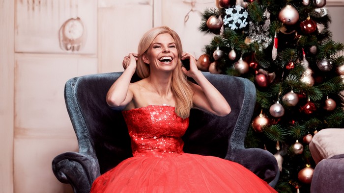 Kvinde i rød kjole sidder i stol nær juletræet.