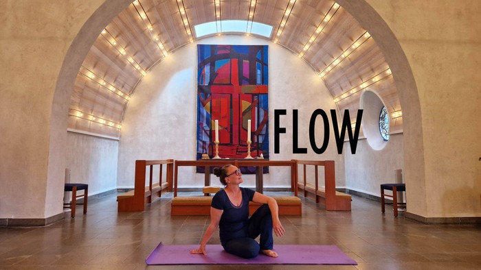 Kvinde i yoga-pose på pink yoga-mat i rum med stor farverig vinduesglasning og ordet