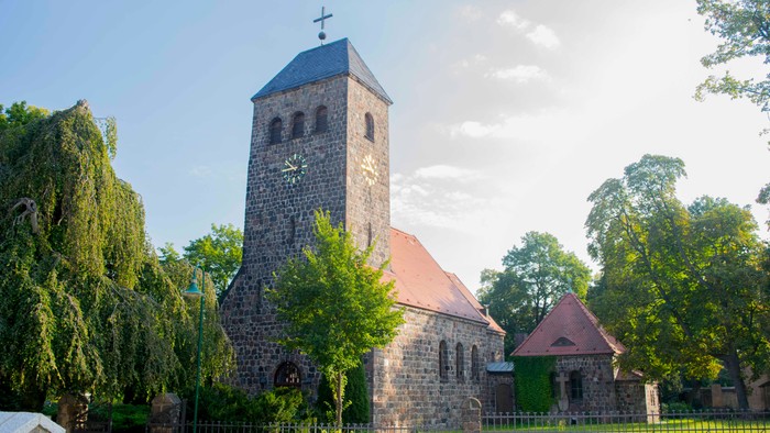Alte Kirche mit hohem Turm und Kreuz, umgeben von Bäumen und einem Zaun