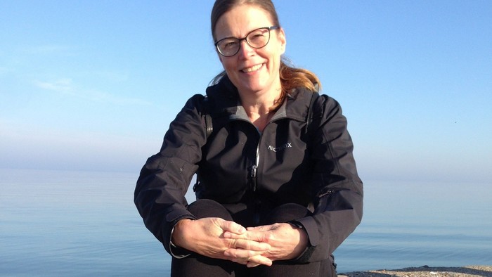 En person sidder på et klippefyldt terræn ved havet iført briller, en sort jakke og sportssko.