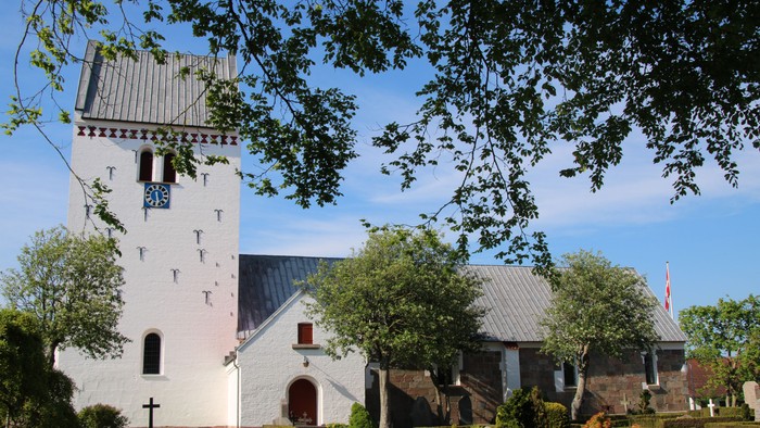 Et hvidt kirkebygning med tårn og blå tak, omgivet af træer og gravsteder