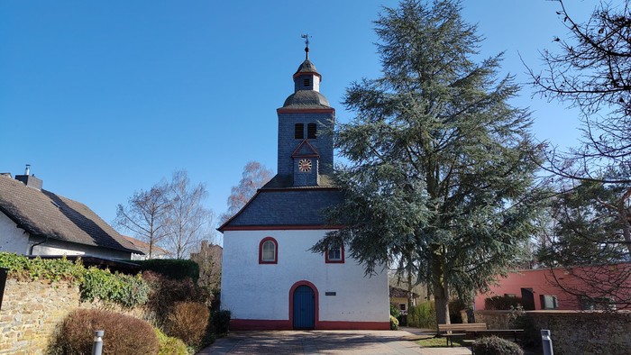 Idyllische weiße Kirche mit Glockenturm in einer ruhigen, landschaftlich gestalteten Dorfgegend.