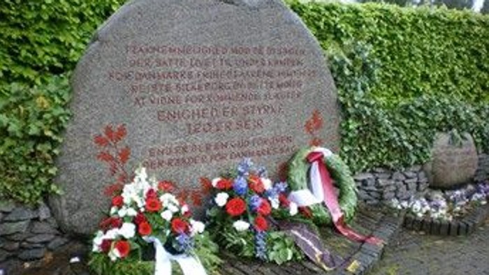 Krigsmindesten prydet med blomsterkranse og indskrevet tekst i en haveindstilling.