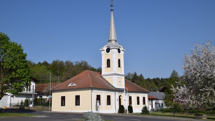 Kleine Kirche mit hohem Turm und Uhr, umgeben von Bäumen und Büschen