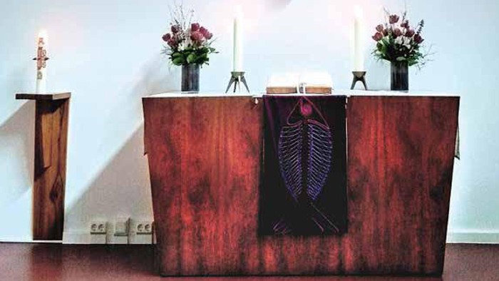 Ein hölzernes Kreuz an der Wand über einem Altar mit Kerzen und Blumen.